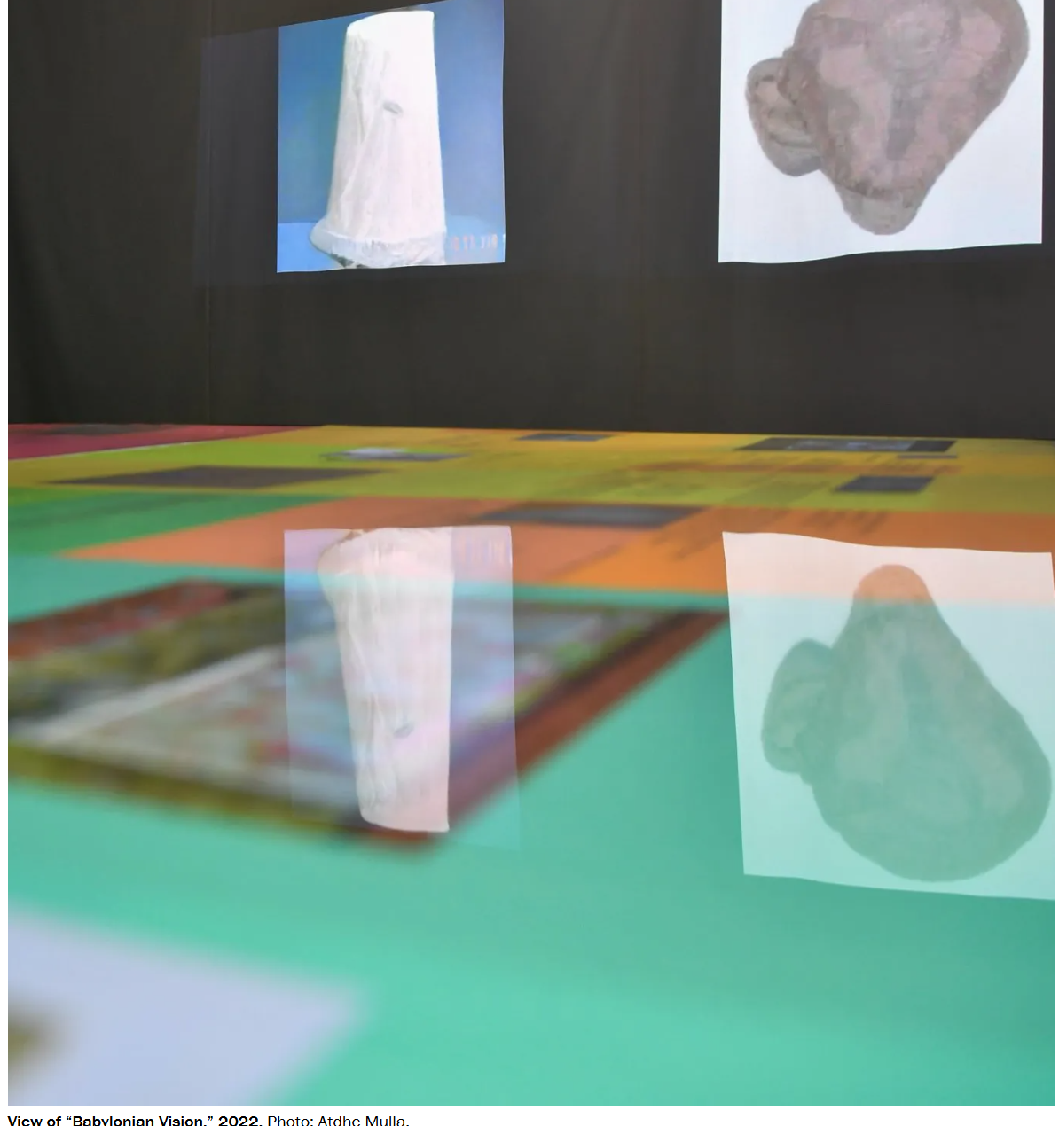 Installation view of Babylonian Vision, 2022: generated artifact images projected on a dark wall and reflected on a green and orange floor