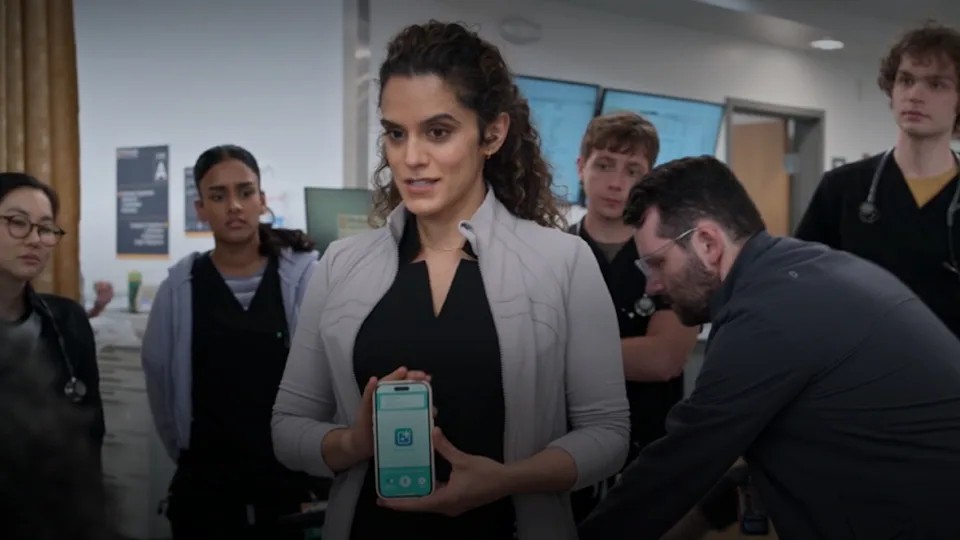 A doctor in The Pitt holds up a phone running an AI charting app while residents look on in the emergency department