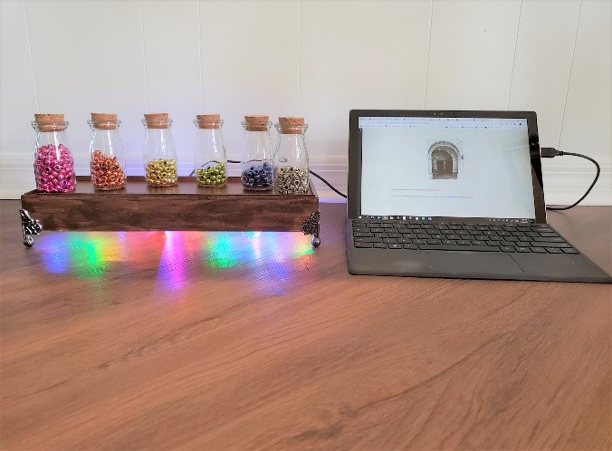 BeadED Adventures: six corked glass jars of beads (pink, orange, gold, green, blue, silver) on a dark wooden stand lit from underneath in rainbow colors, wired to a laptop showing the game.