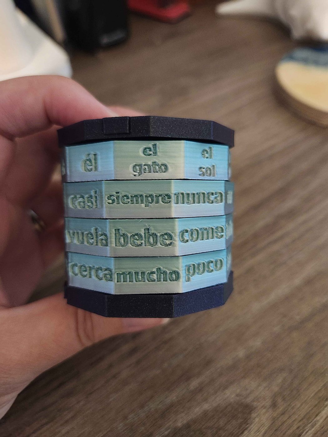 A hand holds Emily’s finished 3D-printed cryptex: pale blue-green octagonal rings between dark navy end caps, each face carrying a recessed Spanish word, reading across “el gato / siempre / bebe / mucho.”