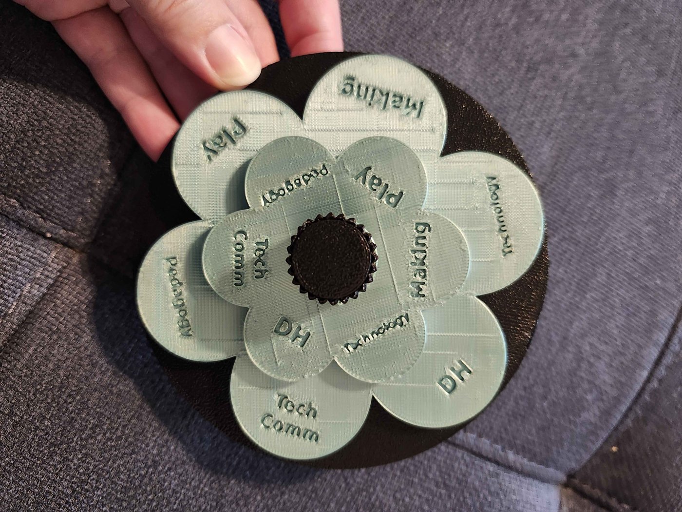A hand holds Emily’s finished 3D-printed flower spinner: two layers of pale green round petals on a black base with a black knob in the center, each petal carrying a recessed word: Making, Play, Technology, DH, Tech Comm, Pedagogy.
