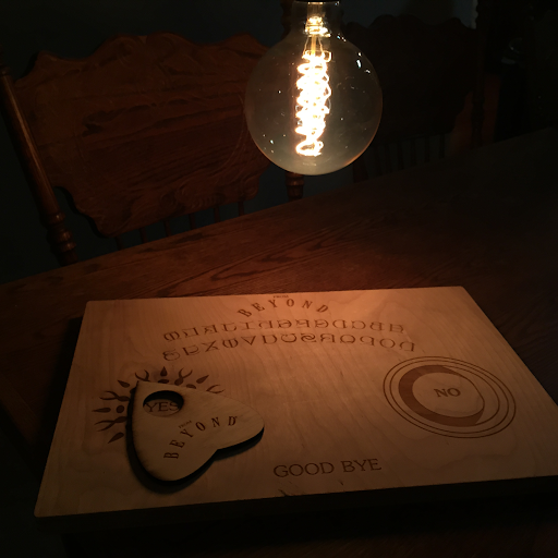 From Beyond: a laser-cut wooden Ouija board reading BEYOND, with a mirrored alphabet, YES, NO and GOOD BYE, and a heart-shaped planchette, lit by a bare filament bulb in a dark room.
